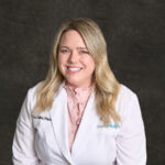 A professional headshot of Tiffany Collins, a blonde woman with a friendly smile, wearing a white pharmacist's coat over a pink ruffled blouse.