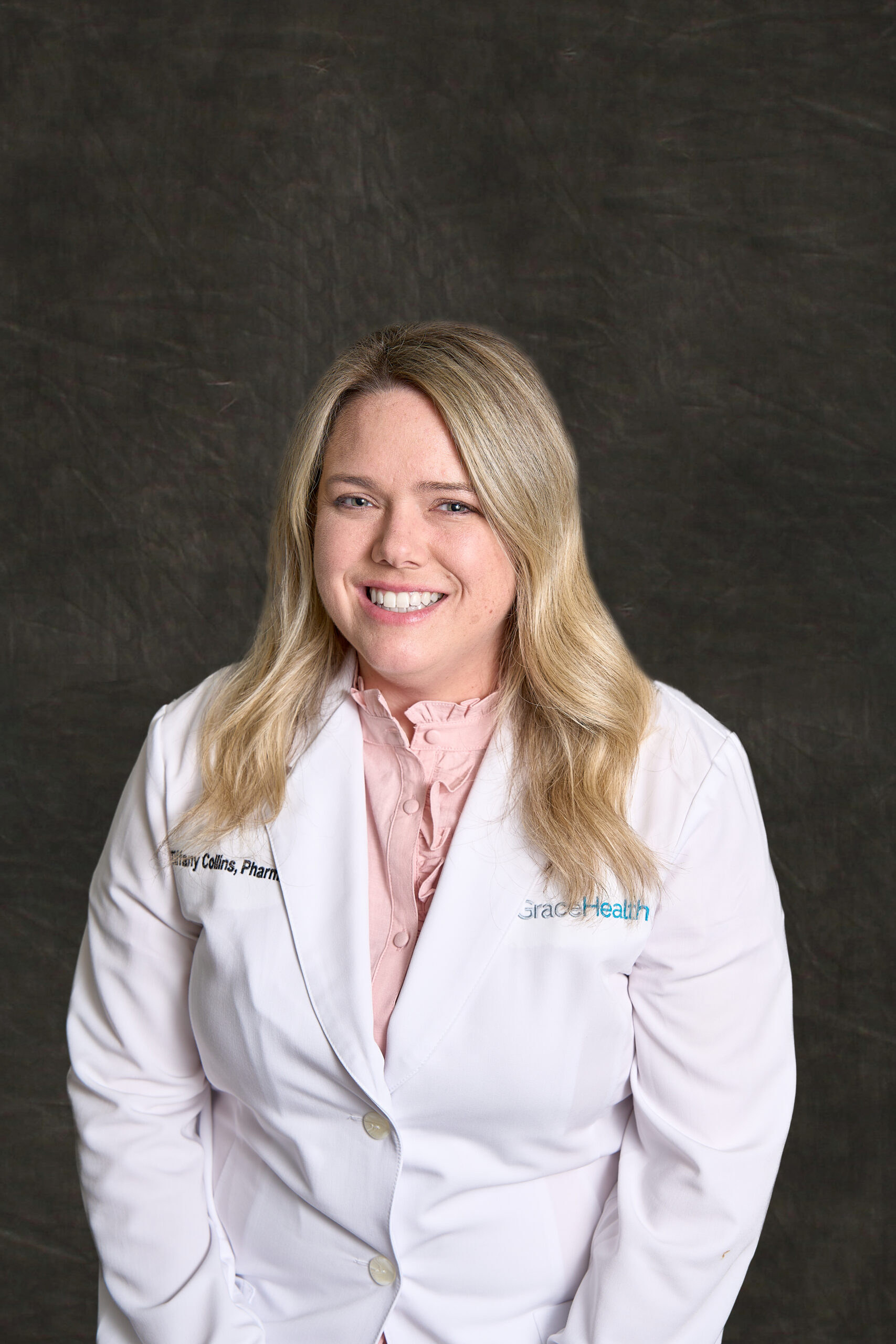 A professional headshot of Tiffany Collins, a blonde woman with a friendly smile, wearing a white pharmacist's coat over a pink ruffled blouse.