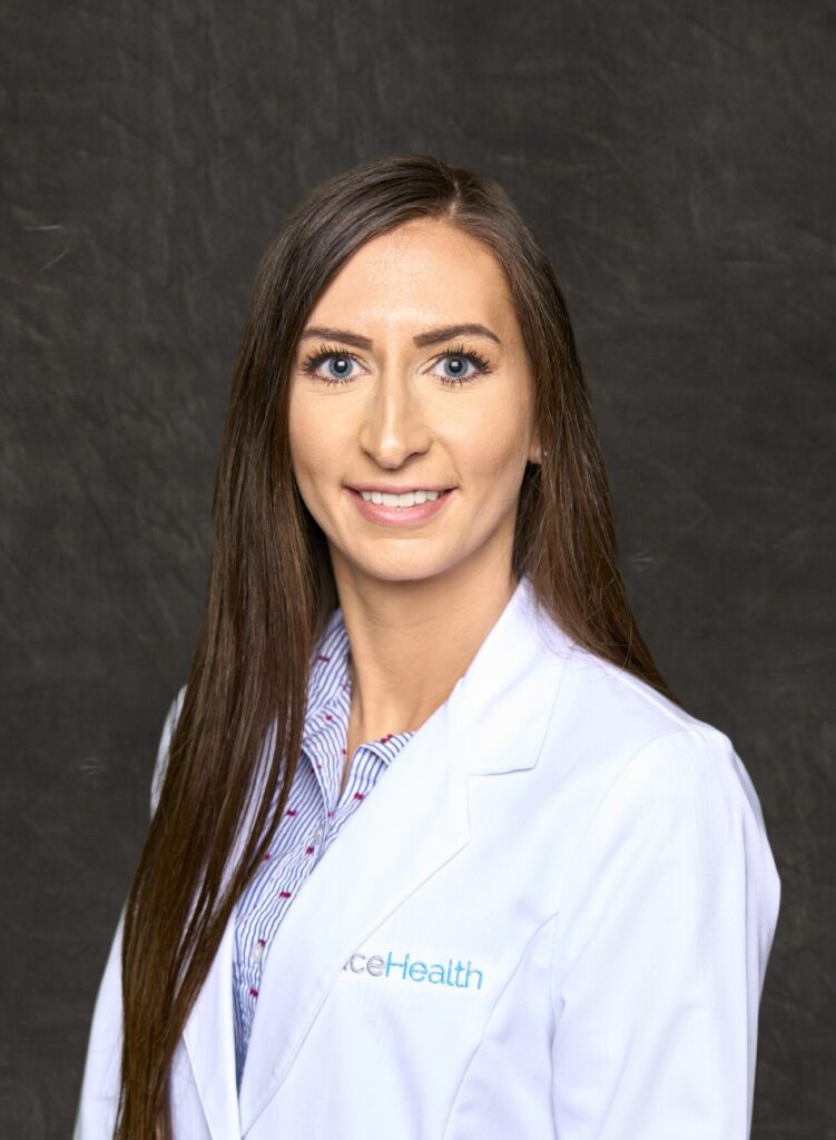A professional headshot of Brittany Grega, a brunette woman with a friendly smile, wearing a white medical coat.