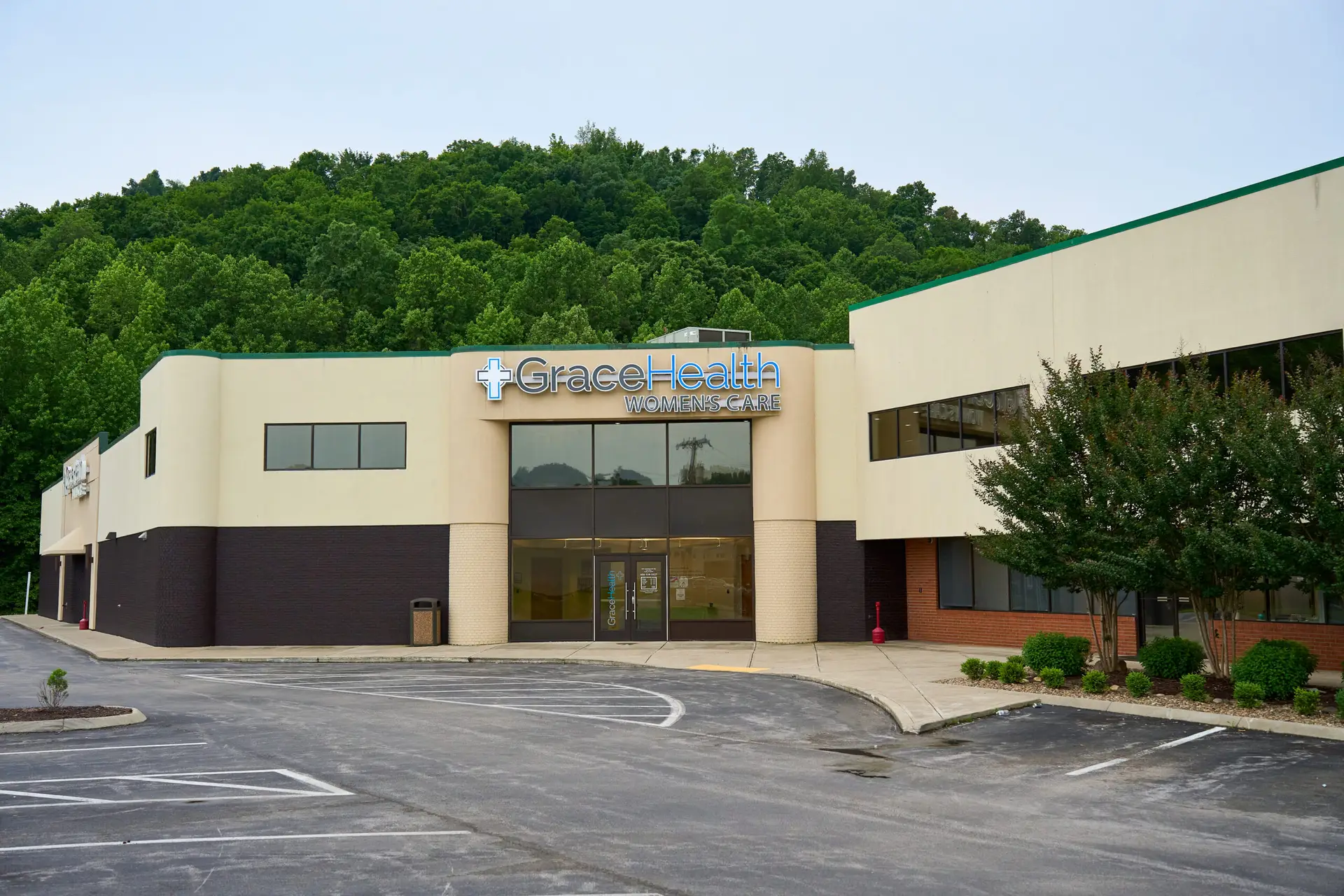 The exterior of the Grace Health Women's Care building, with green trees on a hillside behind it and green shrubbery in front under a light blue sky.
