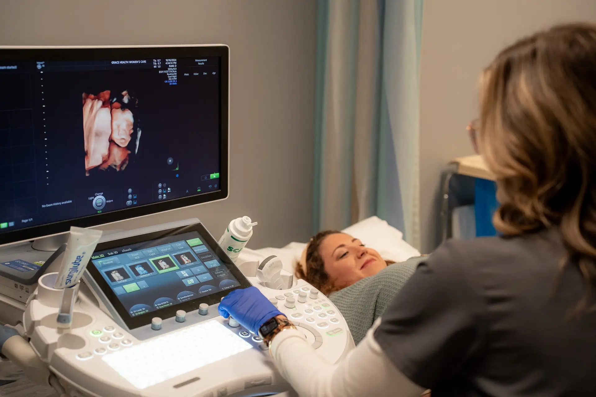 A Grace Health Women's Care patient with dark red hair and a light green shirt smiles happily at an ultrasound screen showing a 3D baby scan; a blurred ultrasound tech in gray scrubs is on the right.