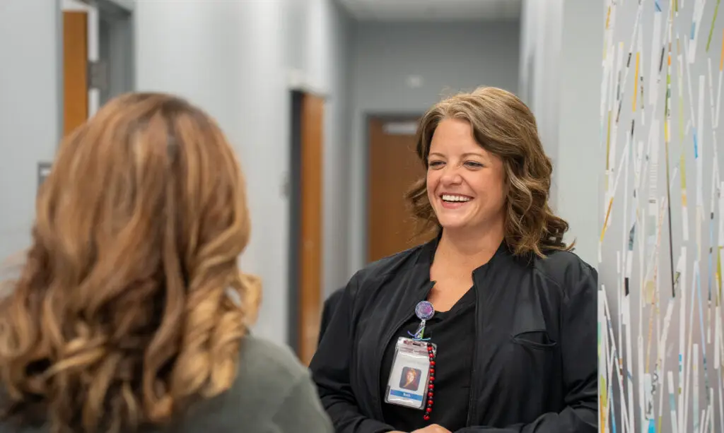 Beth Howard, APRN, in black scrubs, smiles warmly at a subtly blurred coworker in the foreground within the bright, modern clinic of Grace Health Women's Care.