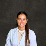 A headshot of Stephanie Beckett, an Advanced Practice Registered Nurse (APRN) at Grace Health. She is a white woman with dark brown hair, brown eyes, and a friendly smile. She is wearing a white lab coat over a beige shirt that has a white border around the neck. The background is a soft, mottled gray.