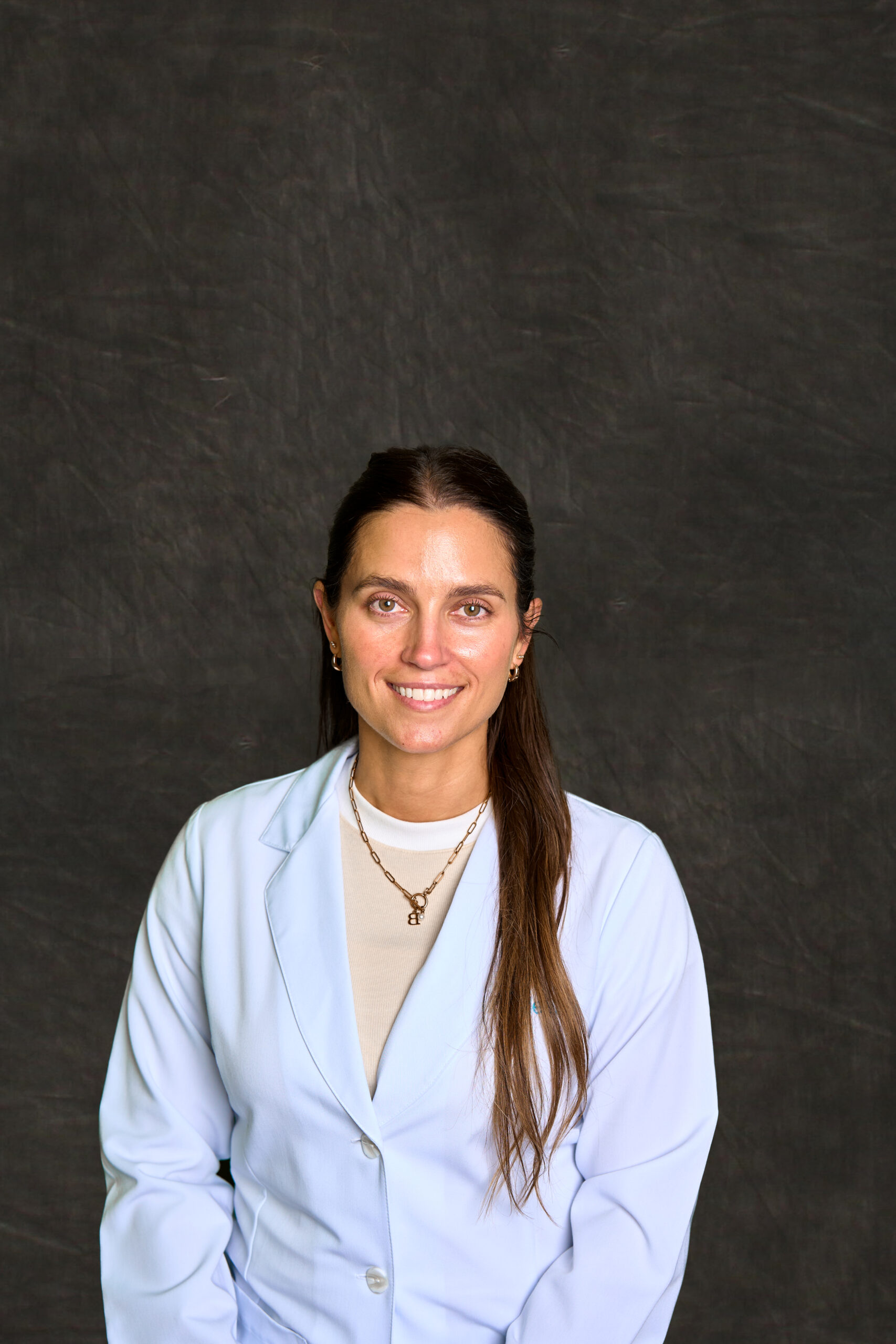 A headshot of Stephanie Beckett, an Advanced Practice Registered Nurse (APRN) at Grace Health. She is a white woman with dark brown hair, brown eyes, and a friendly smile. She is wearing a white lab coat over a beige shirt that has a white border around the neck. The background is a soft, mottled gray.