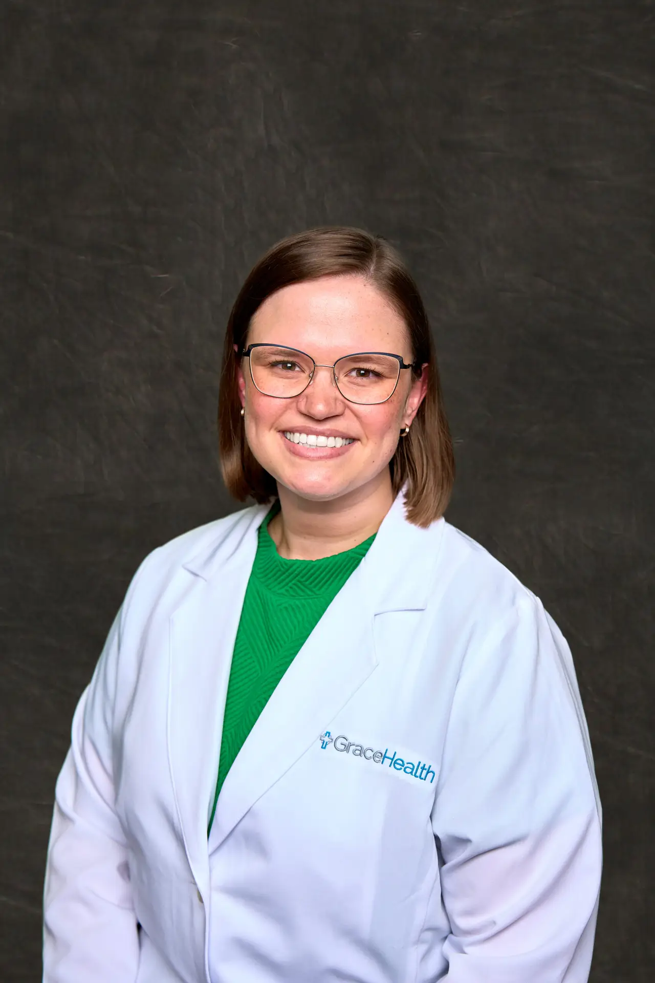 A headshot of Cassidy Pearce, a Physician Assistant at GraceHealth. She is a white woman with brown hair and glasses, wearing a white lab coat with the GraceHealth logo and a green top. She is smiling directly at the camera. The background is a dark, mottled gray.